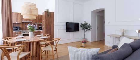 A modern living room with a grey sofa, a wooden dining table with chairs, a flat-screen TV mounted on the wall, and a kitchen area in the background.