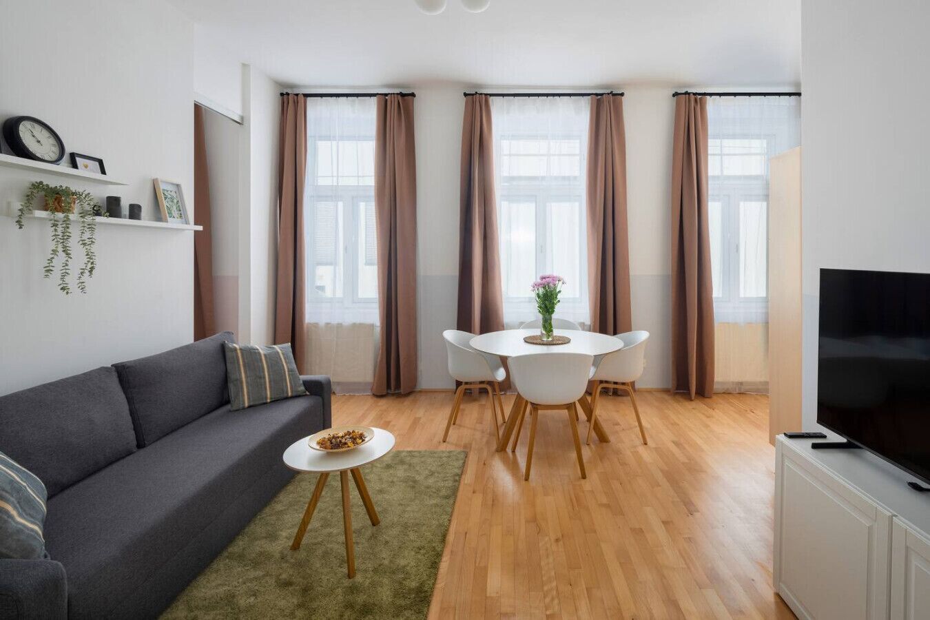 Una sala de estar moderna con un sofá, una mesa de comedor con sillas, un televisor y ventanas grandes con cortinas.