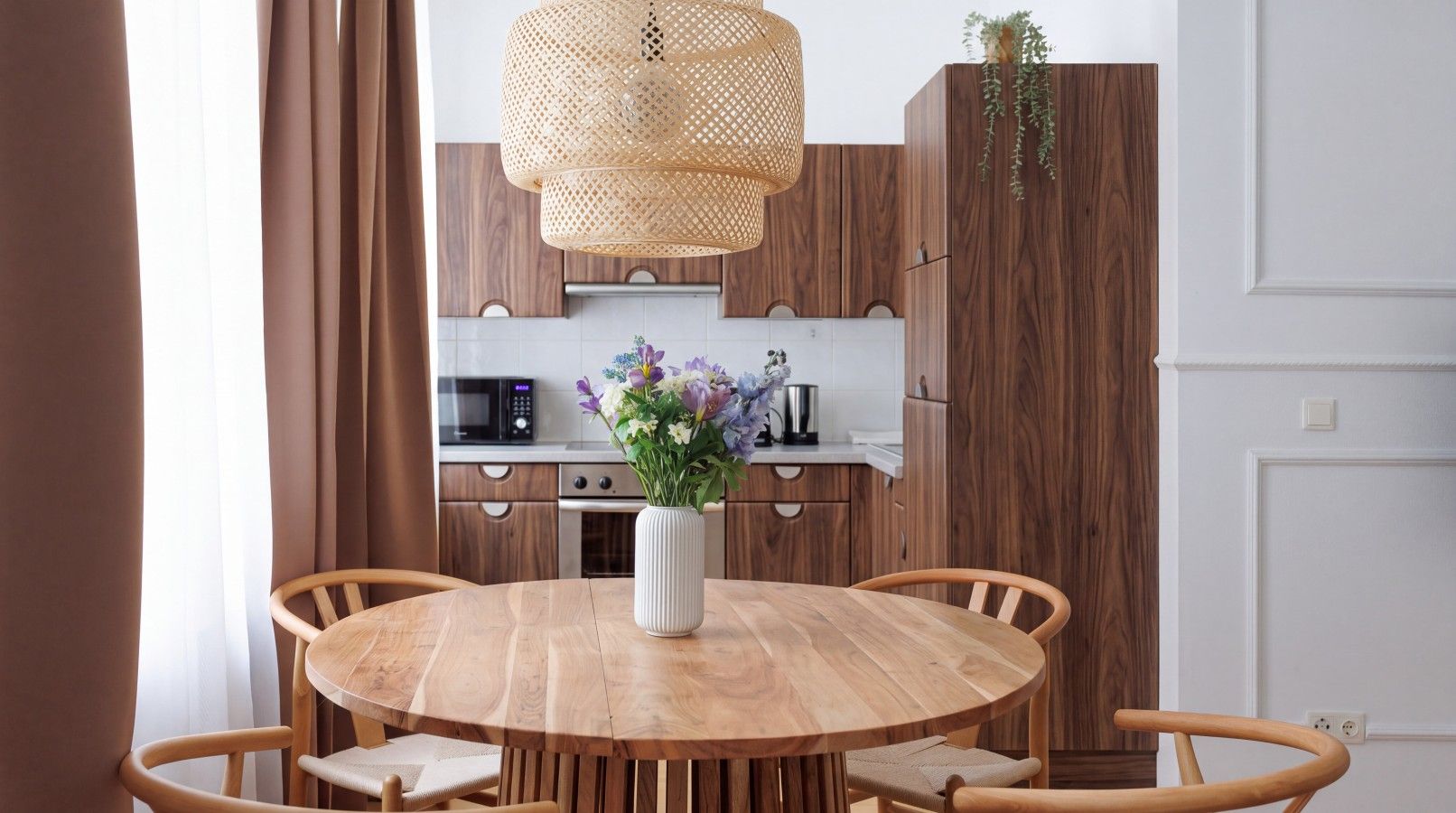 Runder Holz-Esstisch mit vier Stühlen und Blumenstrauß, im Hintergrund die Küchenzeile mit Backofen und Mikrowelle in der Unterkunft in Wien-Ottakring