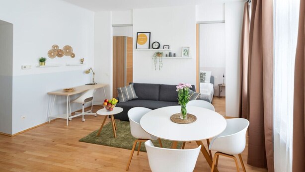 Una sala moderna con una mesa de comedor redonda, sillas blancas, un sofá gris y una pequeña mesita con un cuenco de fruta.