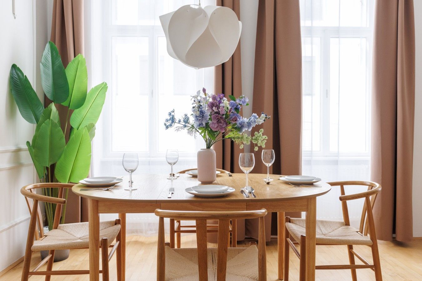 A dining area with a round wooden table, six chairs, a vase with flowers, and a hanging light fixture.