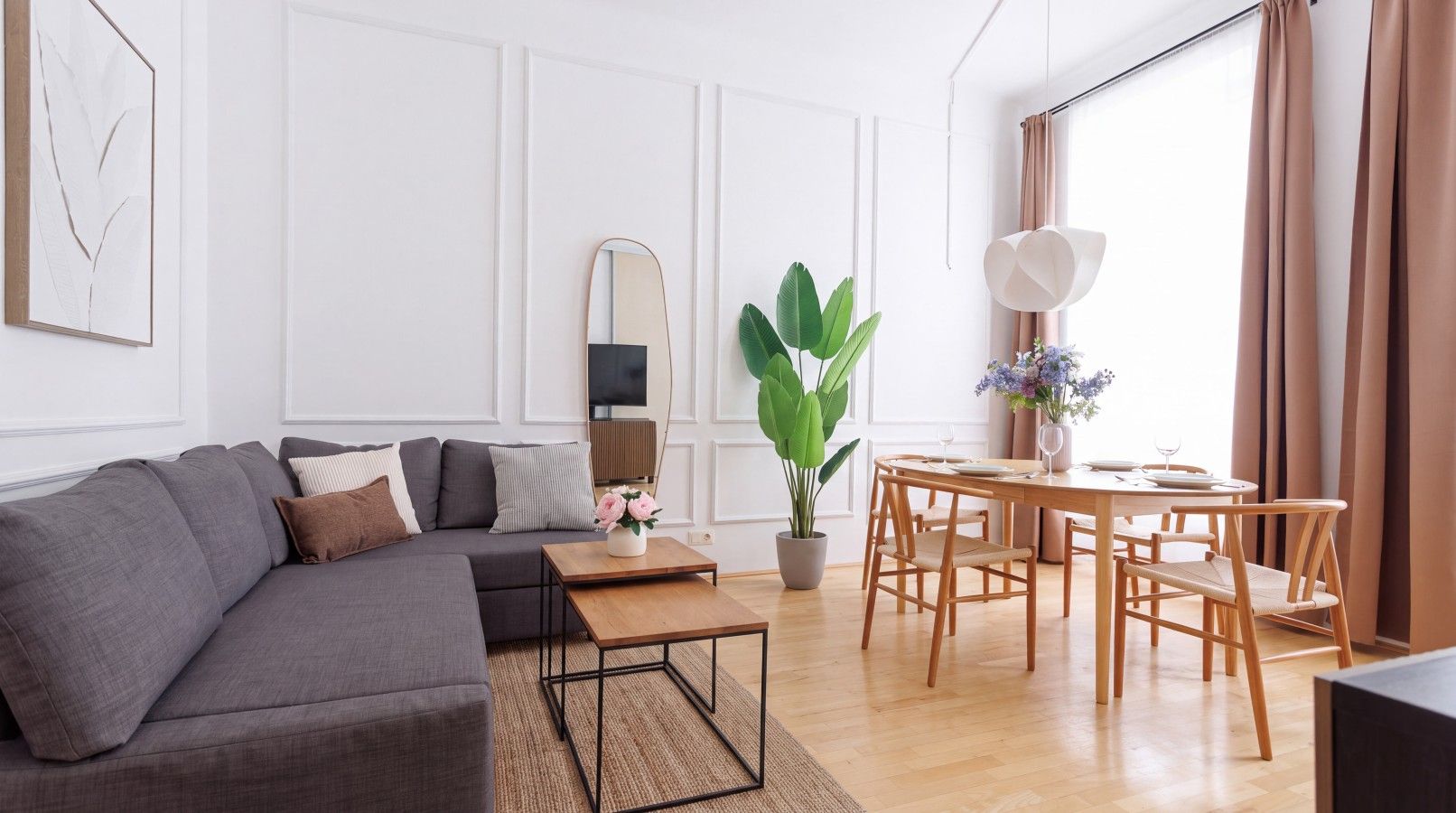 A modern living room with a grey sofa, a wooden coffee table, a dining table with chairs, a large green plant, and a mirror on the wall.