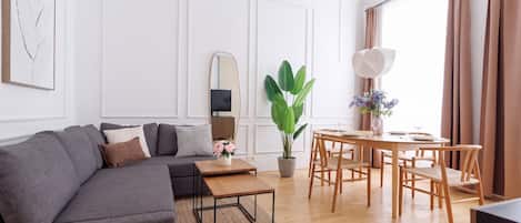 A modern living room with a grey sofa, a wooden coffee table, a dining table with chairs, a large green plant, and a mirror on the wall.