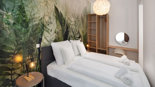 Uma cama com lençóis brancos, um espelho redondo, uma mesinha de cabeceira de madeira e uma parede com um mural de folhagens.