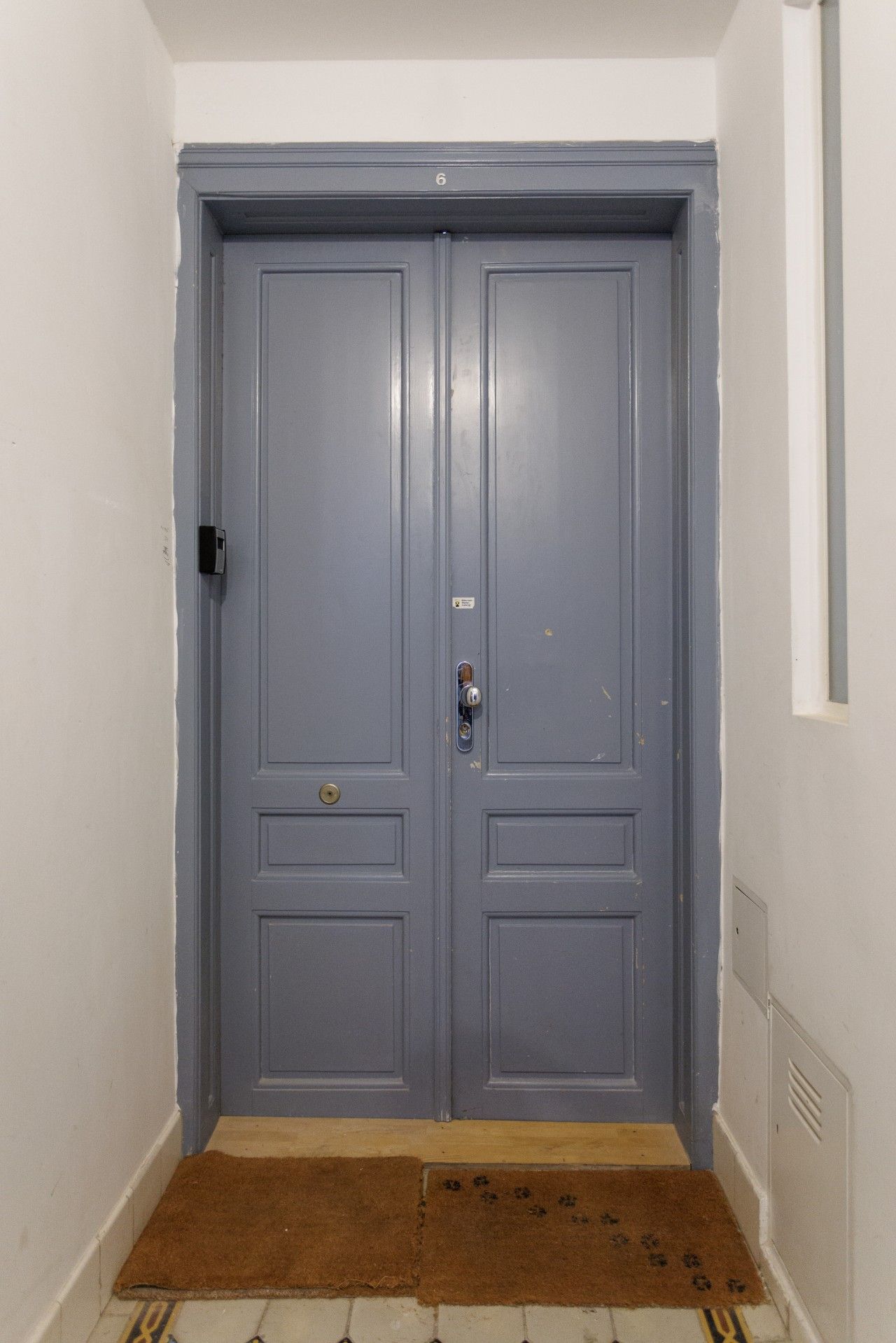 Une porte double fermée, dotée d’une poignée de porte et d’un trou de serrure, un paillasson marron et un mur blanc avec une grille d’aération.