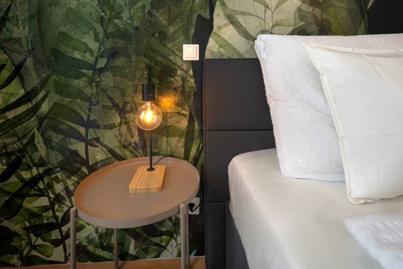 A bed with white bedding, a round bedside table with a lamp, and a wall with a leafy pattern.