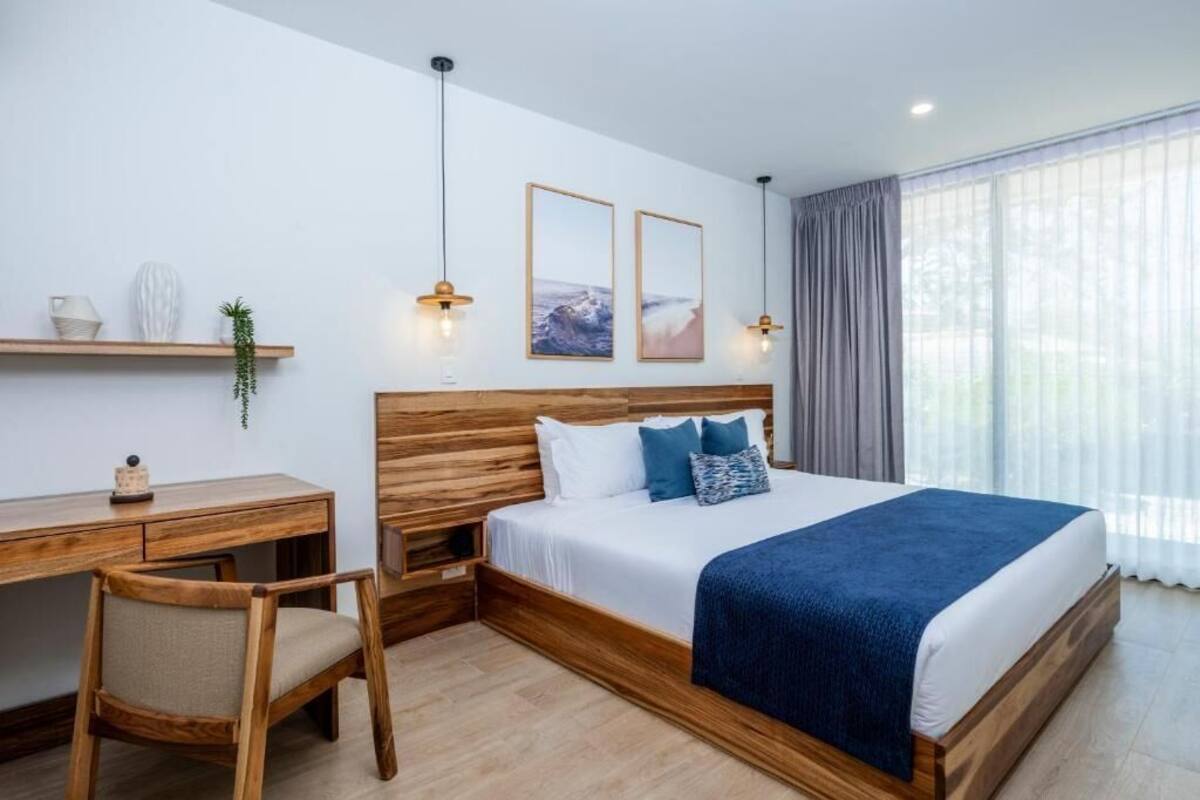 A bedroom with a wooden headboard, a bed with a blue blanket, a desk with a chair, and a window with curtains.