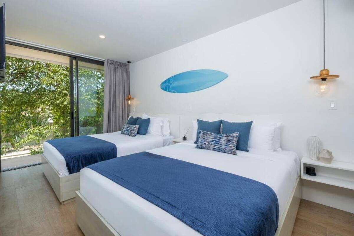 A modern hotel room with two beds, a large window, and a wall-mounted surfboard decoration.