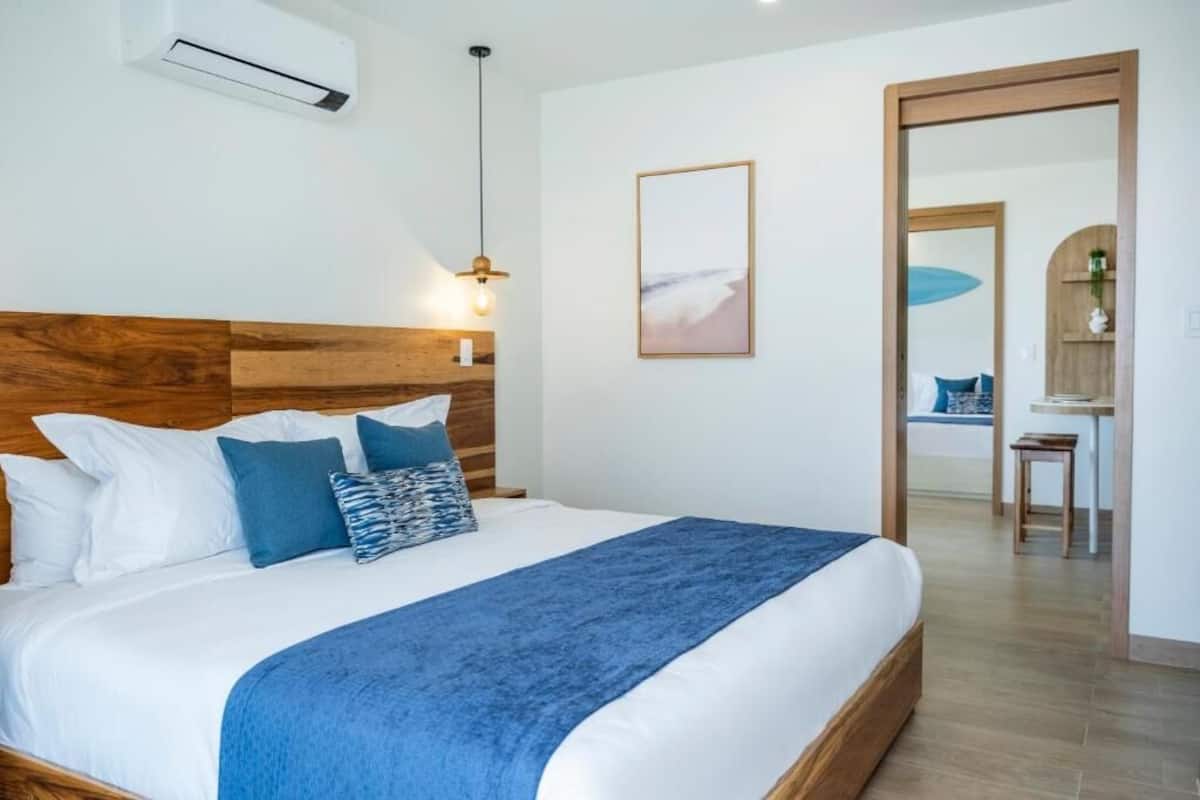 A bedroom with a wooden headboard, a bed with white and blue bedding, a blue throw blanket, a wall-mounted air conditioner, a pendant light, and a framed picture on the wall.