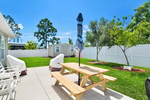 A backyard patio with a wooden table and benches, a covered grill, and a white fence. - Gulf Breeze Coastal Cottage*Beach* Shopping*Dining (Gulf Breeze)