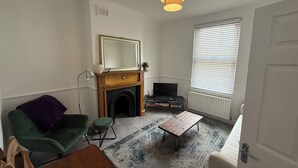 A living room with a fireplace, a television, a green armchair, a wooden coffee table, and a large window with blinds. - Spacious 3 dbl bed house - sleeps 7! On the High St, 10 min walk to East Croydon (Croydon)