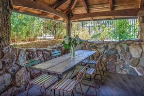 A covered outdoor seating area with a wooden table and metal chairs, surrounded by stone walls and greenery. - Luxury Wine Country Estate Near Downtown/Wineries (Paso Robles)