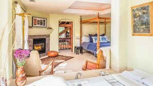 Un dormitorio con cama con dosel, una chimenea, un rincón de estar con una silla y una mesa, y vistas al exterior.