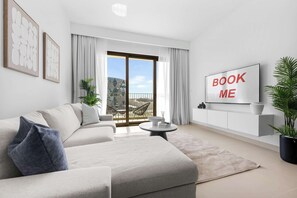 A modern living room with a sofa, a coffee table, a TV, and a balcony with a view. - Free Housekeeping Visits During Your Stay - StayShort - Beautiful Dubai Hills 1BR Apartment that Sleeps Up to 4 (Dubai Hills Estate)