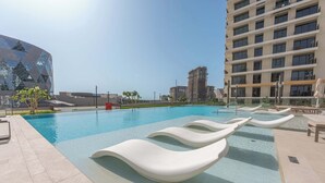 En swimmingpool med liggestole, en moderne bygning og et højt boligkompleks i baggrunden.