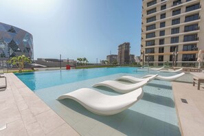 A swimming pool with lounge chairs, a modern building, and a high-rise apartment building in the background. - Free Housekeeping Visits During Your Stay - StayShort - Beautiful Dubai Hills 1BR Apartment that Sleeps Up to 4 (Dubai Hills Estate)