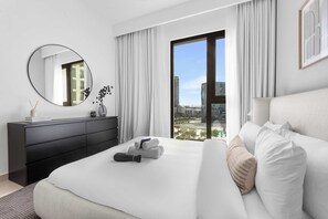 A modern hotel room with a large bed, a black dresser, a round mirror, and a view of the city through a large window. - Free Housekeeping Visits During Your Stay - StayShort - Beautiful Dubai Hills 1BR Apartment that Sleeps Up to 4 (Dubai Hills Estate)