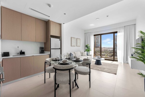 A modern kitchen with wooden cabinets, a dining area with round tables and chairs, and a balcony with a view of the city. - Free Housekeeping Visits During Your Stay - StayShort - Beautiful Dubai Hills 1BR Apartment that Sleeps Up to 4 (Dubai Hills Estate)
