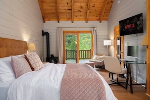 A hotel room with a bed, a desk, a chair, a TV, and a balcony with a view of greenery.