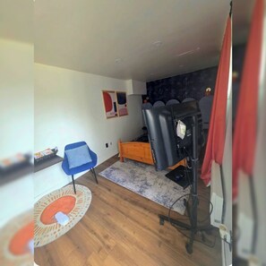 Una habitación con una silla azul, una cama de madera, una silla de masaje negra, una alfombra pequeña y una pared con arte abstracto.