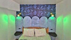 Quarto com cama, cabeceira com iluminação e parede com padrão de folhas.
