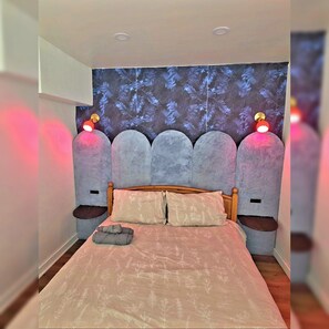 Un dormitorio con cama, cabecera con luces, tapiz en la pared y mesita de noche.