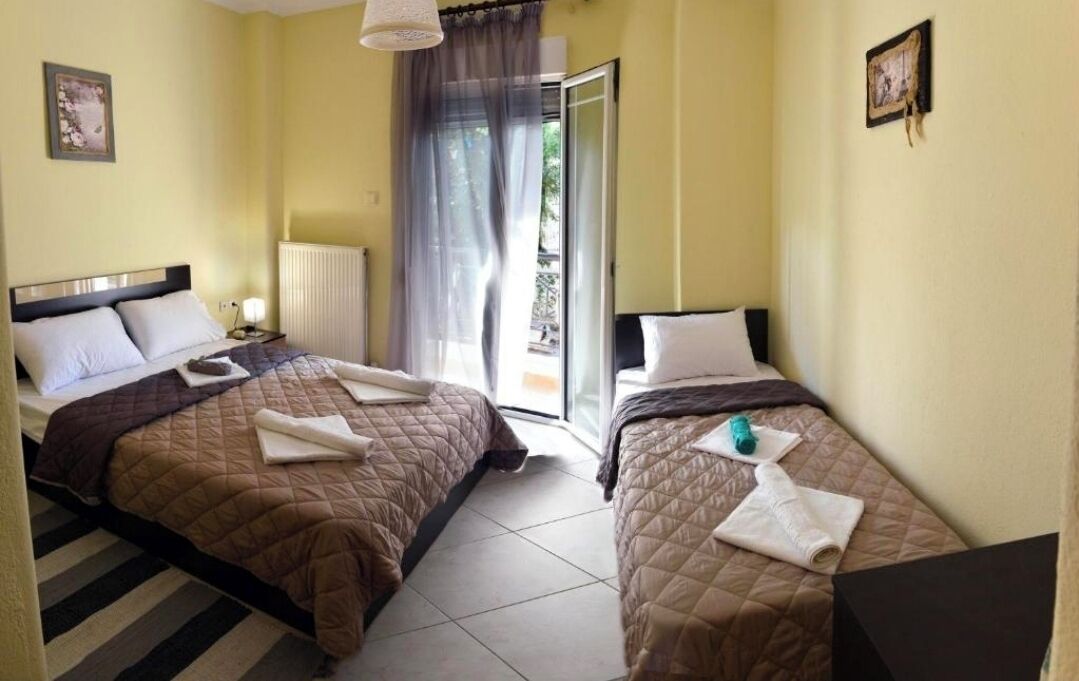 A hotel room with two beds, a radiator, a window with sheer curtains, and a view of greenery outside.