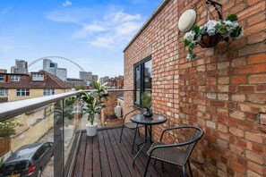 Ein Balkon mit Holzboden, zwei Stühle, einem Tisch, einem hängenden Blumenkorb und Blick auf die Stadt.