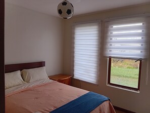 Un dormitorio con cama, almohadas, una mesita de noche, una ventana con persianas y un ventilador de techo.