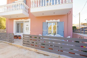 A pink building with a balcony, a white door, and a bench with potted plants. - Nikorina Apts | 1 (Apolpena)