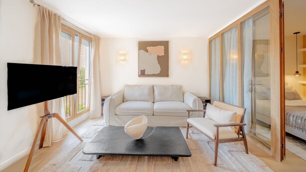 Una sala moderna con una televisión de pantalla plana, un sofá, una silla, una mesa de centro y un puerta de vidrio que da a un dormitorio.