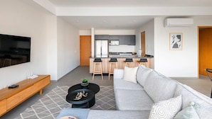 Uma sala de estar moderna com um sofá, uma mesa de centro e uma televisão. Uma cozinha com bancada e banquetas pode ser vista ao fundo.