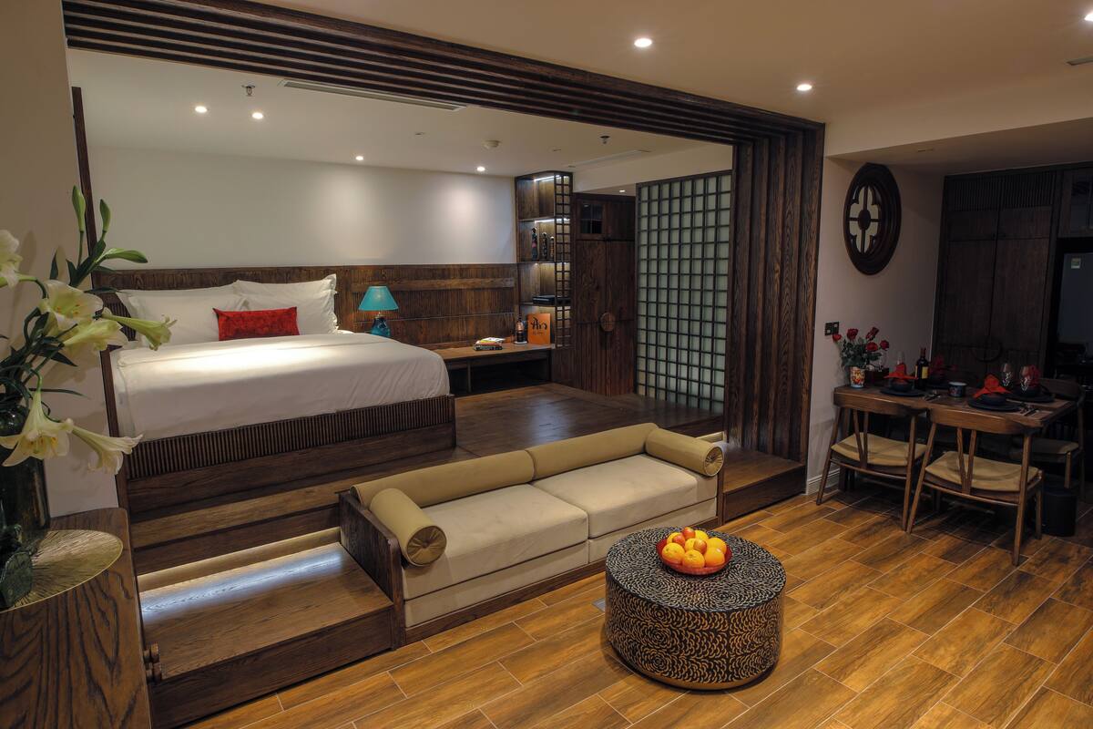 A modern hotel room with a bed, sofa, round coffee table, and a dining area with chairs.