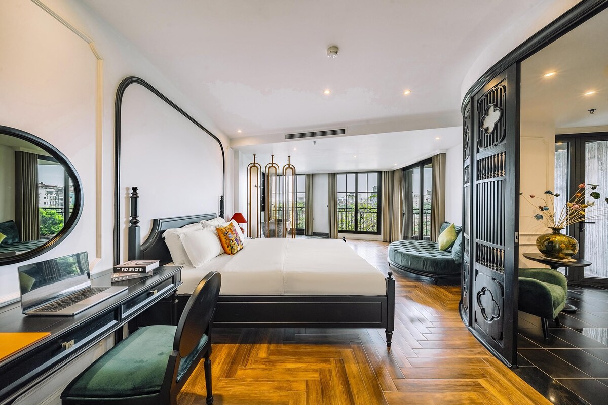 Ein modernes Schlafzimmer mit einem großen Bett, einem Schreibtisch mit Laptop, einem Sessel und Blick auf die Stadt durch große Fenster.
