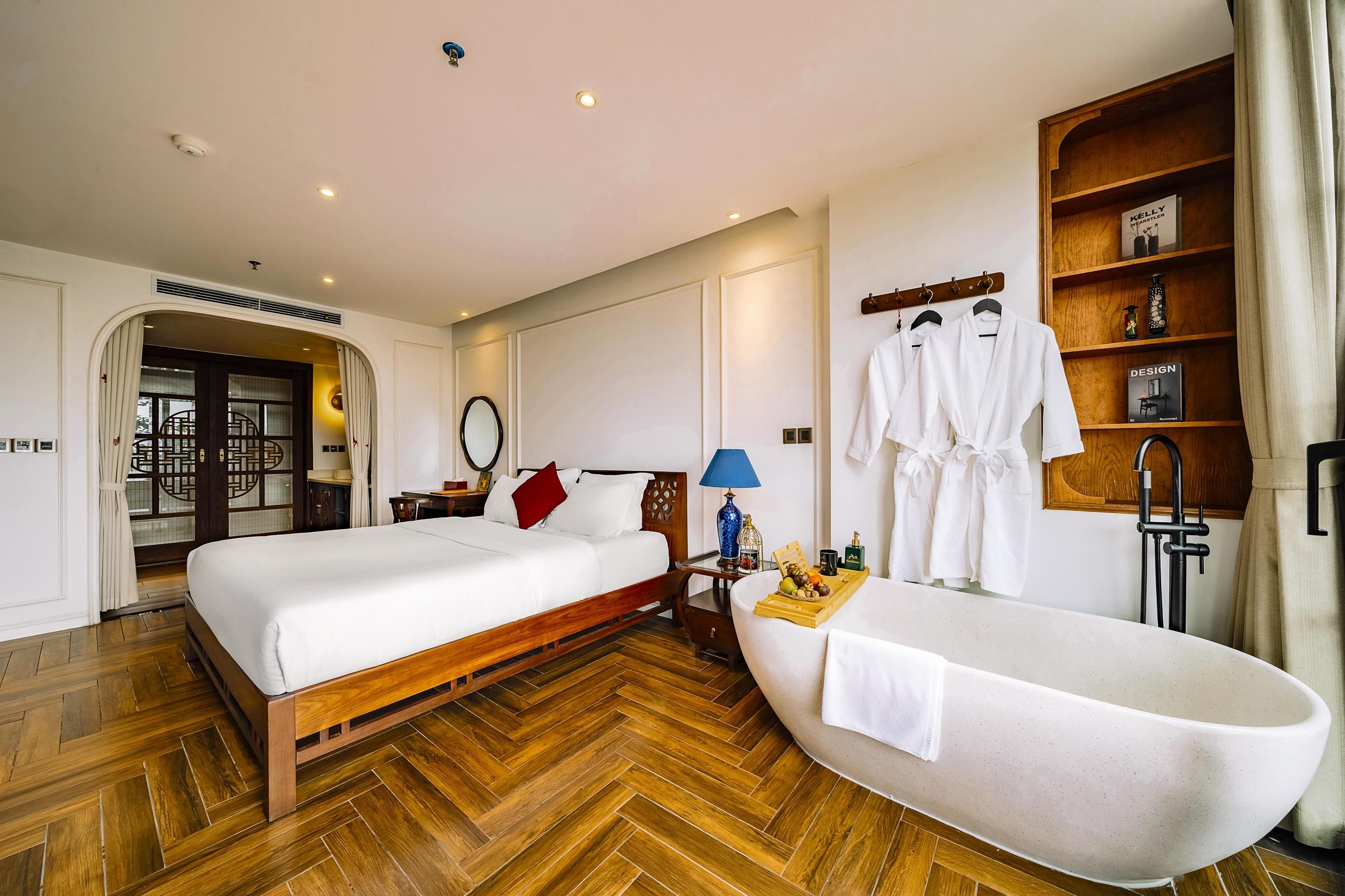 Habitación de hotel con piso de madera en patrón de espiga, una cama grande con ropa blanca, una mesita de noche de madera, una mesita auxiliar con lámpara, un espejo, un estante de madera con batas, y una bañera.
