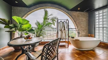 A modern bathroom with a freestanding bathtub, a round table with chairs, and a view through an arched window.
