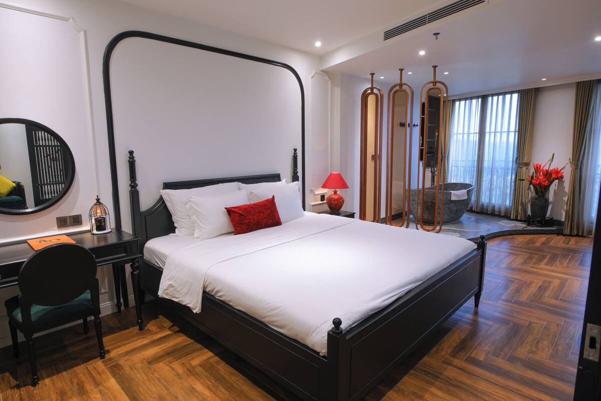 A modern hotel room with a large bed, a desk, a chair, a mirror, and a view of the outdoors.
