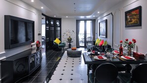 A modern living room with a black and white tiled floor, a large flat-screen TV, a dining area with a glass table, and a seating arrangement with green and yellow cushions.