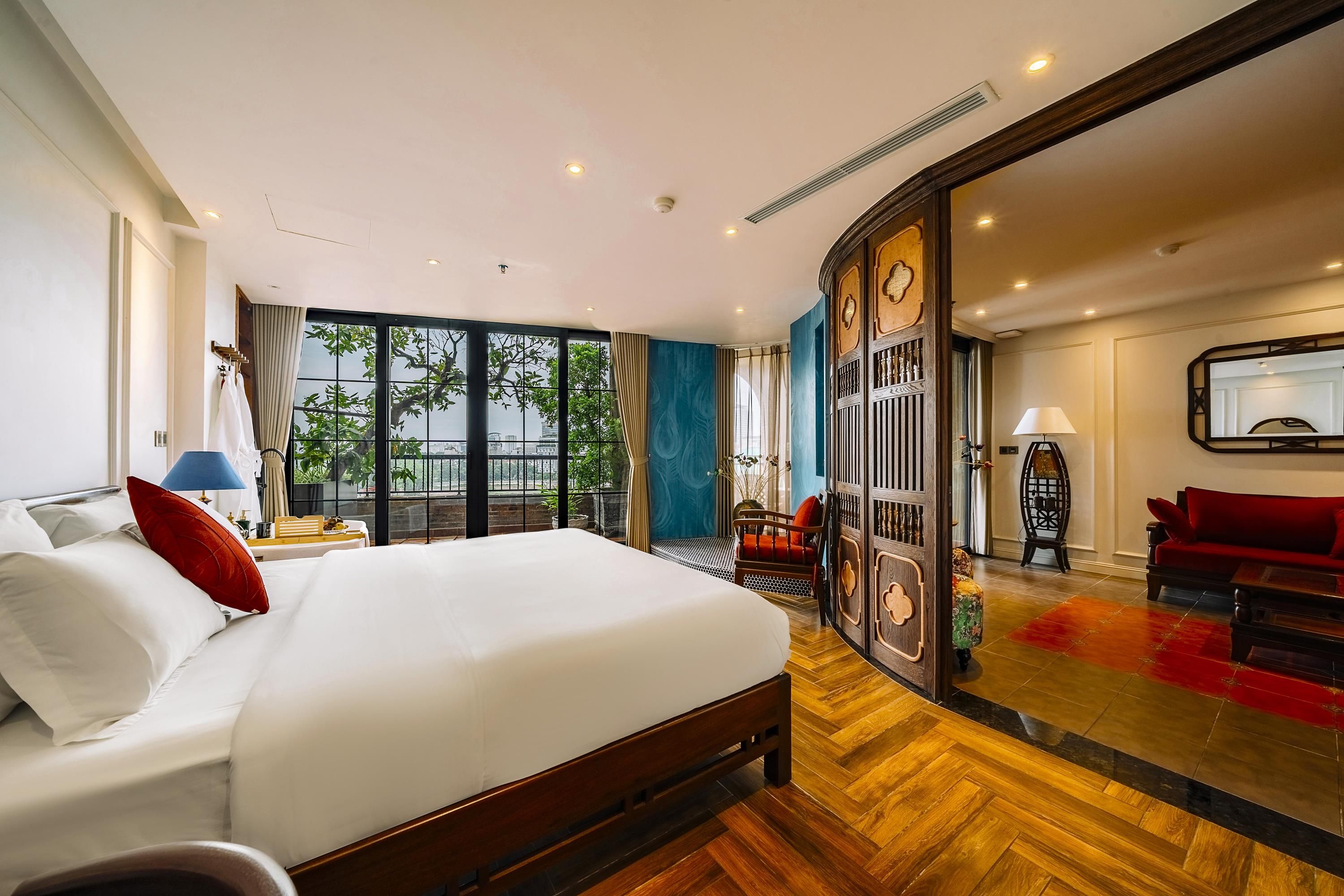 Quarto de hotel com uma cama grande, um espaço de estar com sofá vermelho e vista para a cidade através de grandes janelas.