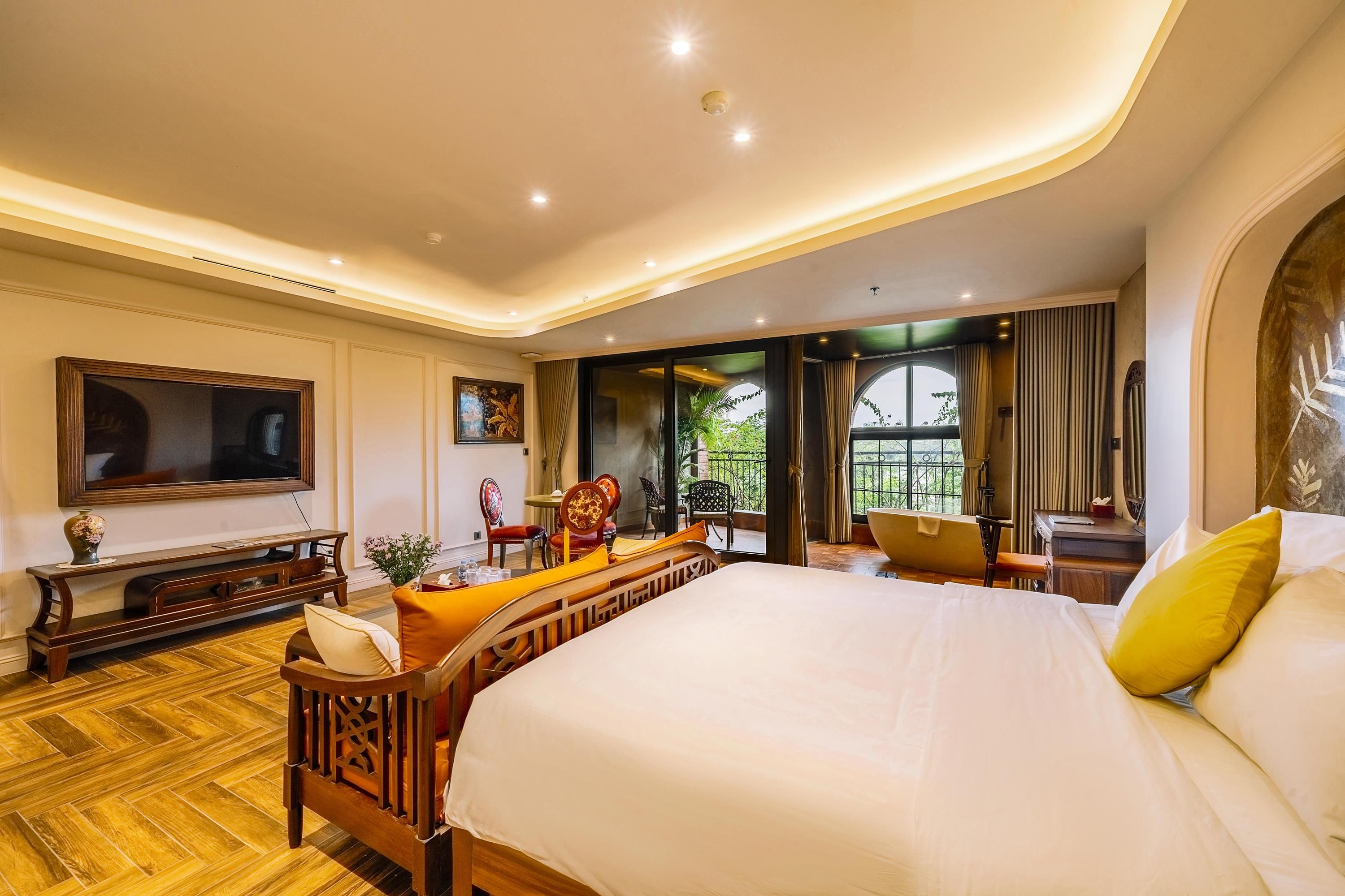 Habitación de hotel con una cama grande, televisión, zona de estar con sillas y vistas a zonas verdes.