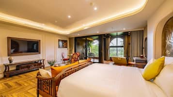 A hotel room with a large bed, a television, a seating area with chairs, and a view of greenery outside.