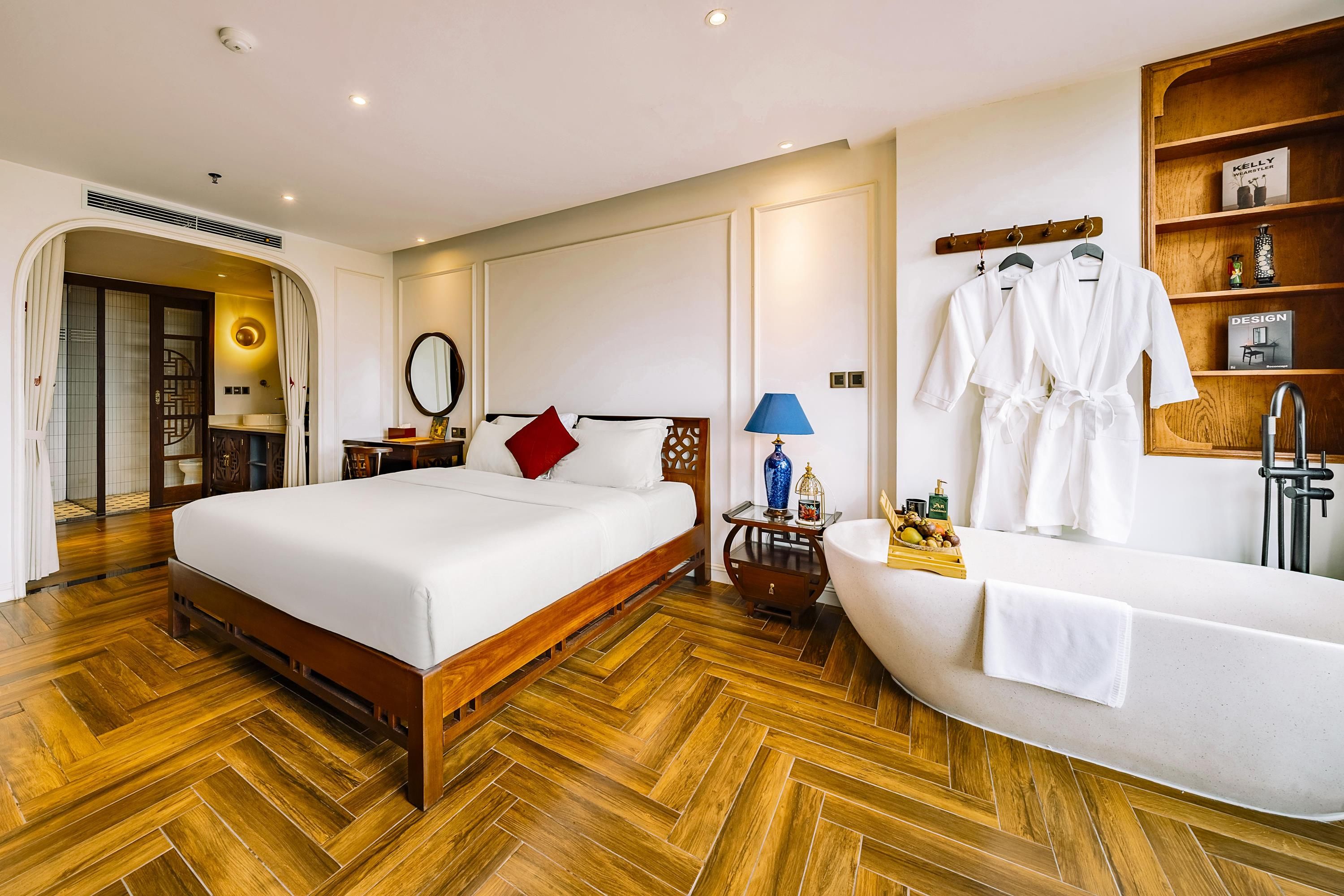 A hotel room with a wooden herringbone floor, a large bed with white bedding, a bedside table with a lamp, a vanity with a mirror, and a bathtub with a towel.