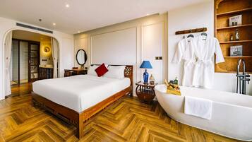A hotel room with a wooden herringbone floor, a large bed with white bedding, a bedside table with a lamp, a vanity with a mirror, and a bathtub with a towel.