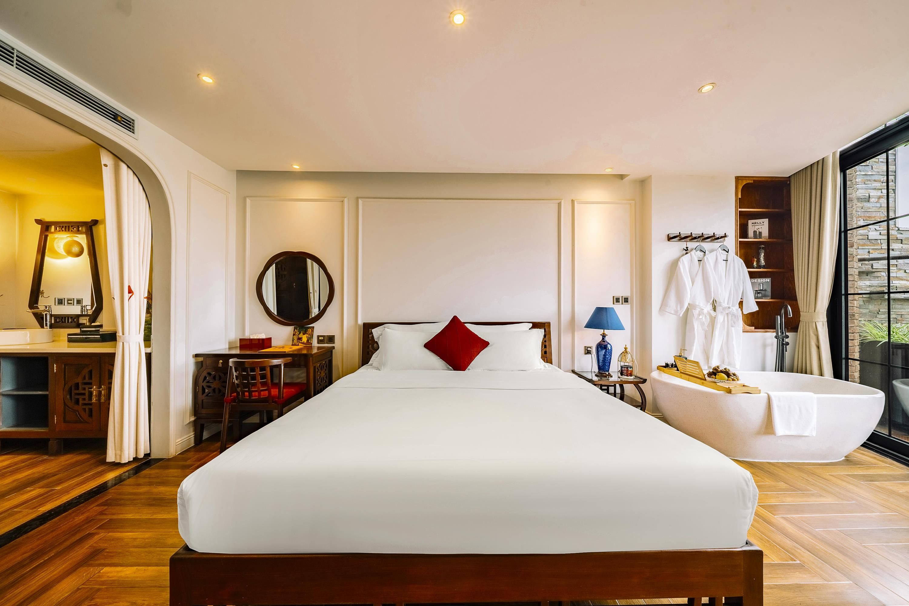 Ein Hotelzimmer mit einem großen Bett, einem Schreibtisch mit Lampe, einem Schminktisch mit Spiegel und Blick ins Freie.