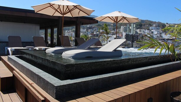 Une piscine à débordement située sur le toit, avec des chaises longues et des parasols, entourée d’une terrasse en bois et offrant une vue sur la ville.