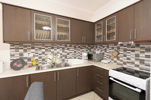 A modern kitchen with dark wooden cabinets, a tiled backsplash, a stainless steel sink, a microwave, and a stove. - Nikorina Apts | 2 (Apolpena)