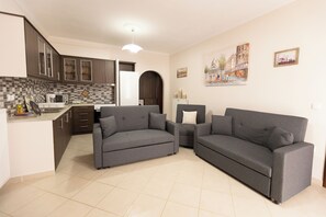 A modern living room with a grey sofa set, a kitchen area with dark cabinets, and a tiled floor. - Nikorina Apts | 2 (Apolpena)