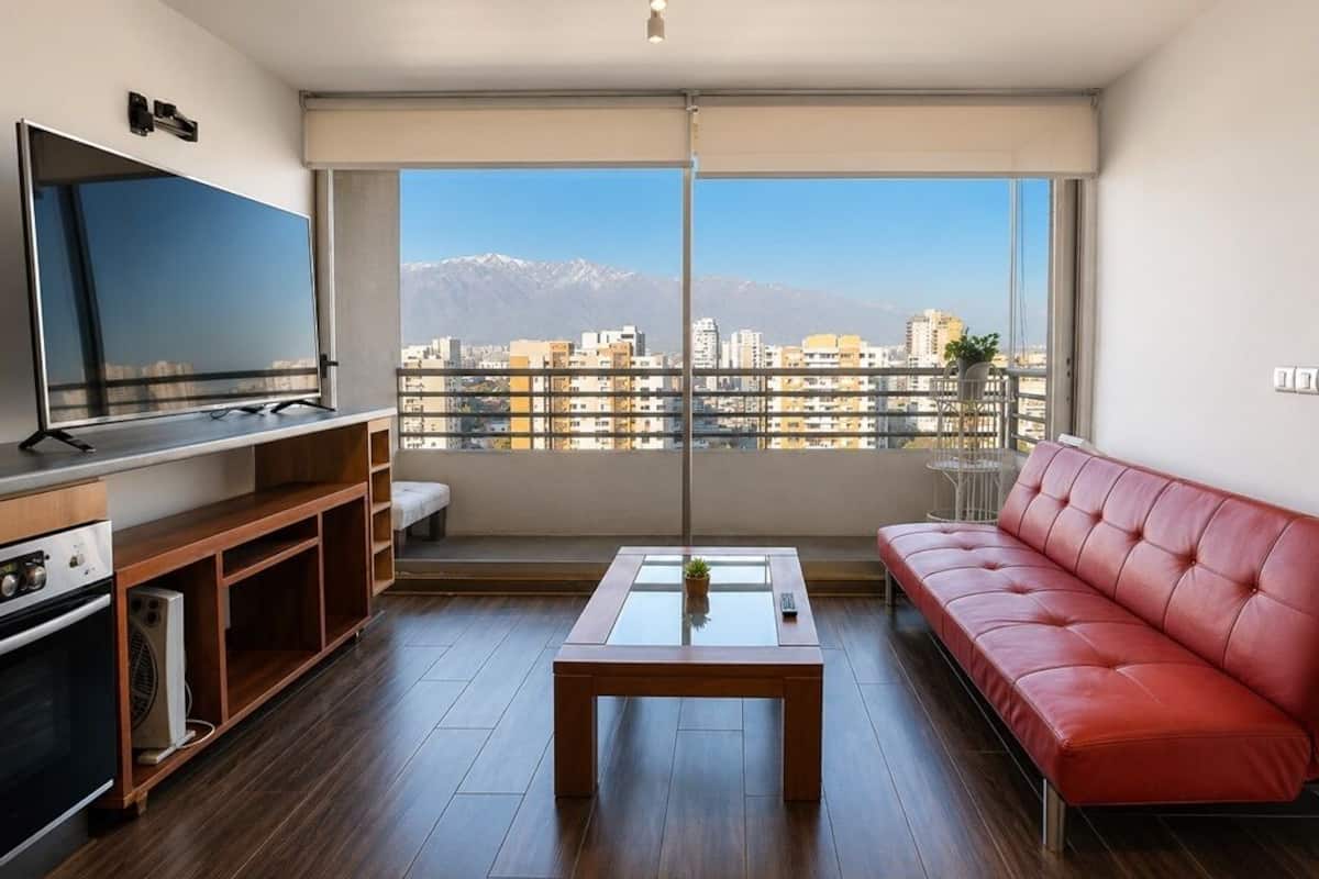 Ett modernt vardagsrum med en stor TV, en röd soffa, ett soffbord i glas och utsikt över en stadssiluett med berg i bakgrunden.