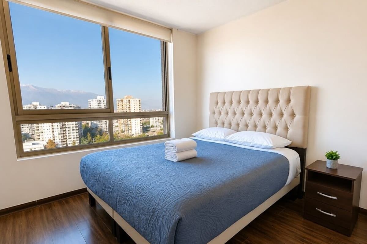A bedroom with a large window, a bed with a blue bedspread, a tufted headboard, and a small bedside table with a plant.