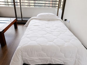 A single bed with a white quilt, a bench, and a window with a view of buildings.
